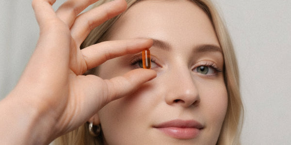 Gros plan sur une jeune femme blonde tenant une capsule rouge nu3 Curcuma Forte entre son pouce et son index devant son œil.