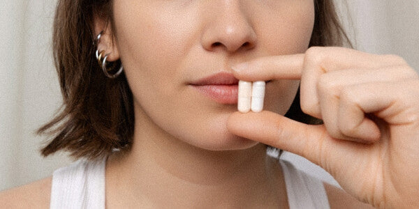 Gros plan sur le visage d'une jeune femme brune tenant deux gélules entre le pouce et l'index à hauteur de la bouche